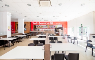 Diferentes mesas para comer y al fondo se observa la entrada al Self Service donde el cliente puede coger su comida